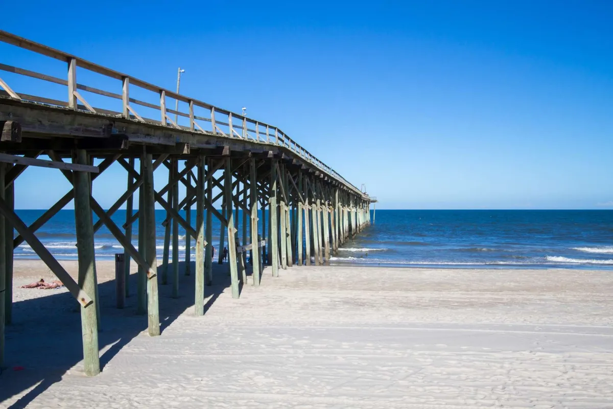 A classic ocean pier stretches into the water with sandy shores in the foreground. The scene is ideal for fishing, walking, or building sandcastles, evoking a sense of seaside tradition and fun for all ages.
