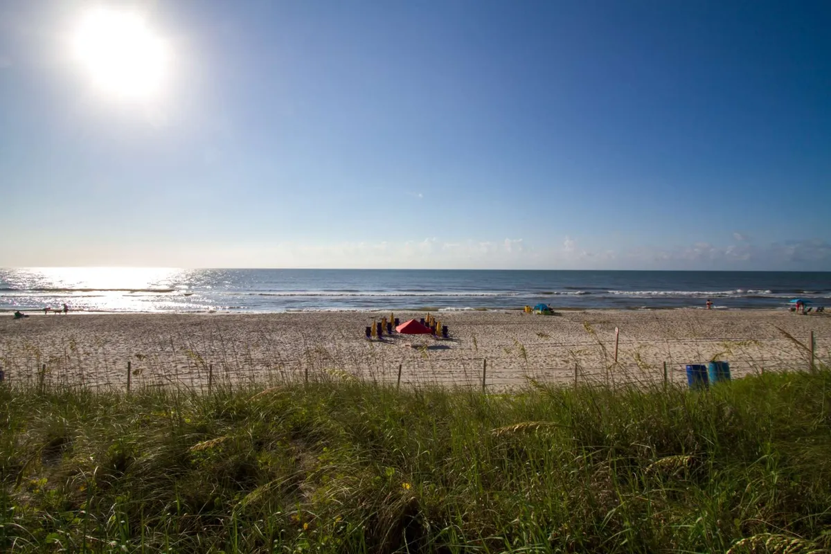 An energetic scene on the beach with the sun shining over the ocean, people gathered under a tent, and others enjoying sunbathing, swimming, or playing games. The mood is cheerful and social, highlighting group fun and beachside camaraderie.