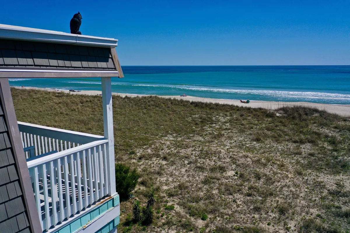 A peaceful exterior view from a deck overlooking the ocean and sandy beach. The deck’s railing features prominently, with a seagull perched nearby and gentle waves meeting the shore. The panorama is ideal for observing sunrises or soaking up the coastal atmosphere while working or relaxing.