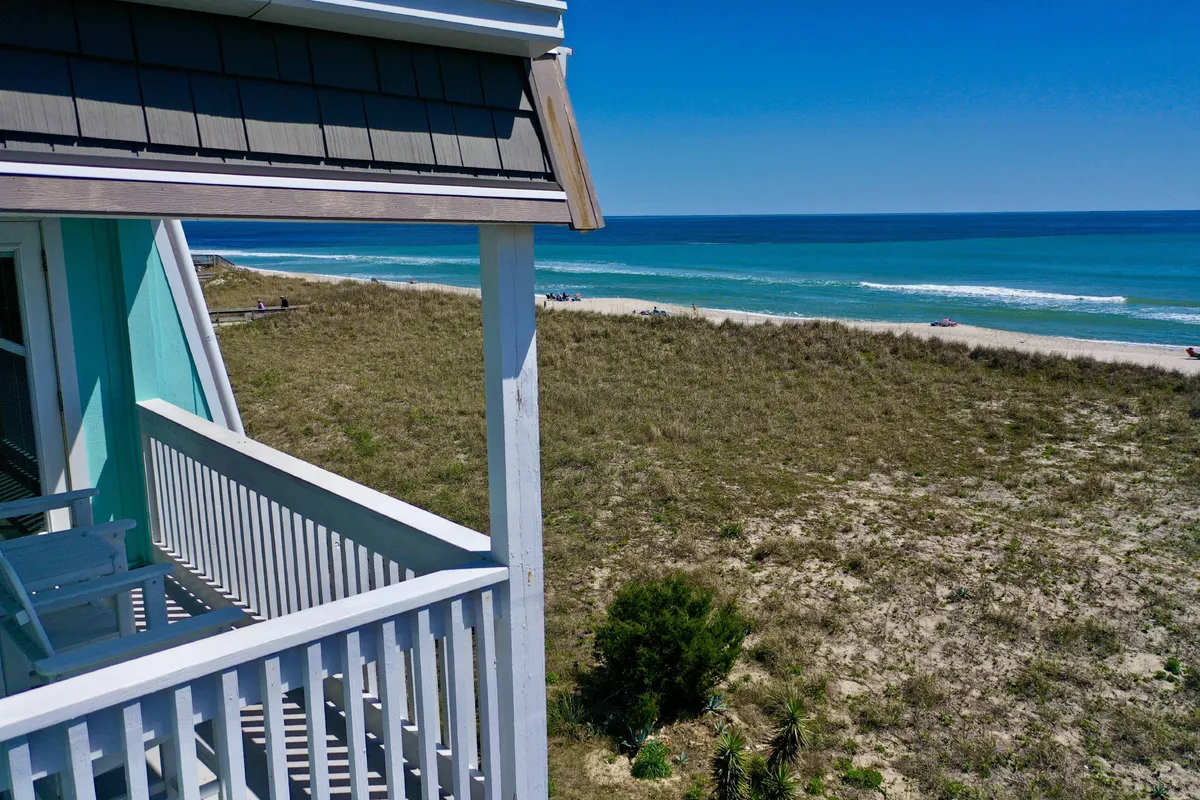 A serene balcony view overlooking rolling ocean waves, sandy beach, and lush greenery. The vantage point from the deck invites quiet moments, with the tranquil scene epitomizing the private oasis described for morning coffees or evening wines.