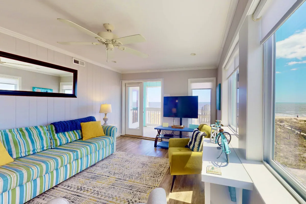 A bright, open-plan living room featuring a striped sofa and large windows that overlook the ocean. The space is cheerful, with casual decor, a TV for movie nights, and a decorative bicycle adding personality. Natural light floods the room, emphasizing the panoramic ocean views and inviting atmosphere.