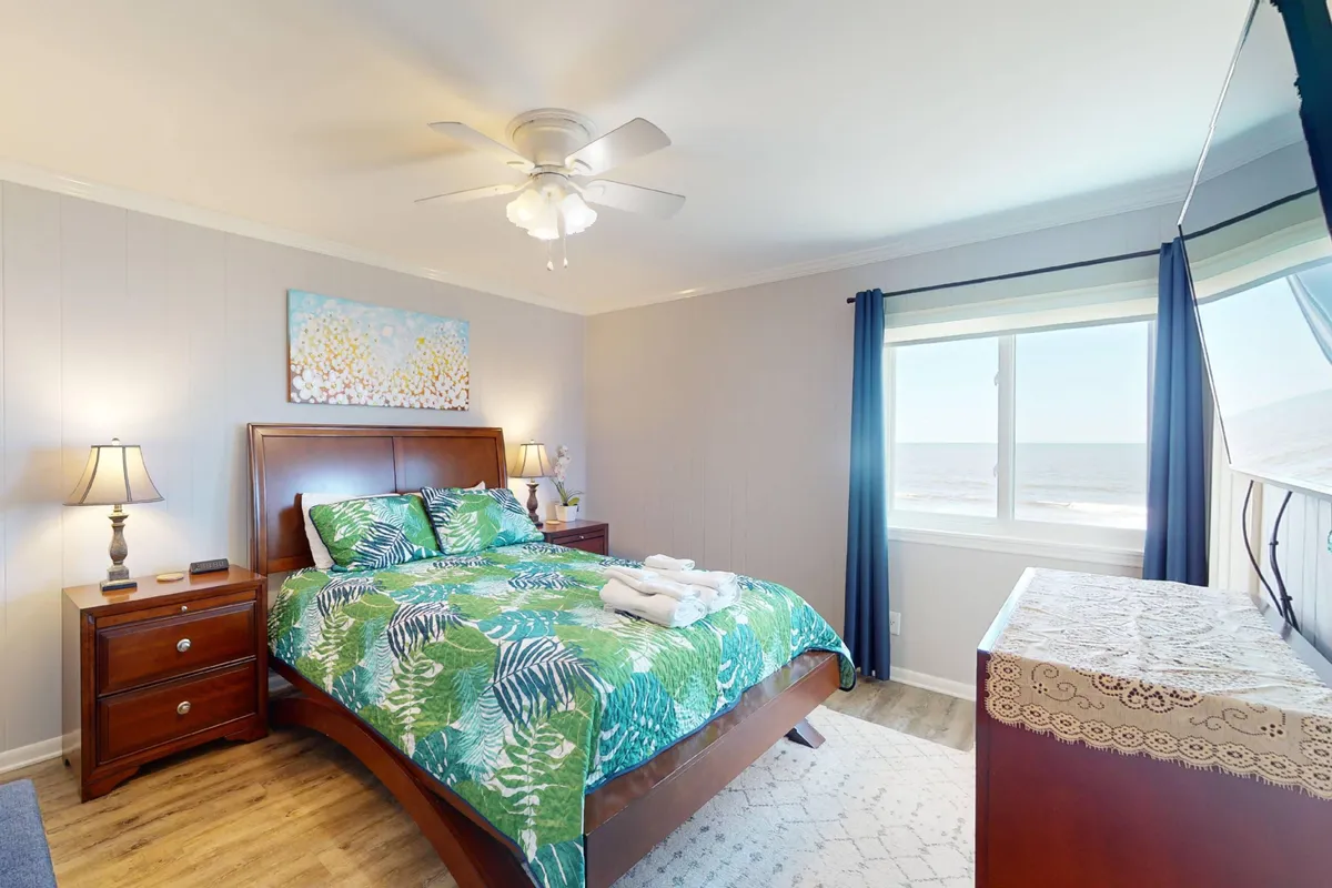 This image features a queen bed with tropical bedding, a large window offering an ocean view, a nightstand with a lamp, and a decorative painting on the wall. The space feels airy and tranquil, with natural light streaming in to create a calming atmosphere ideal for rest.
