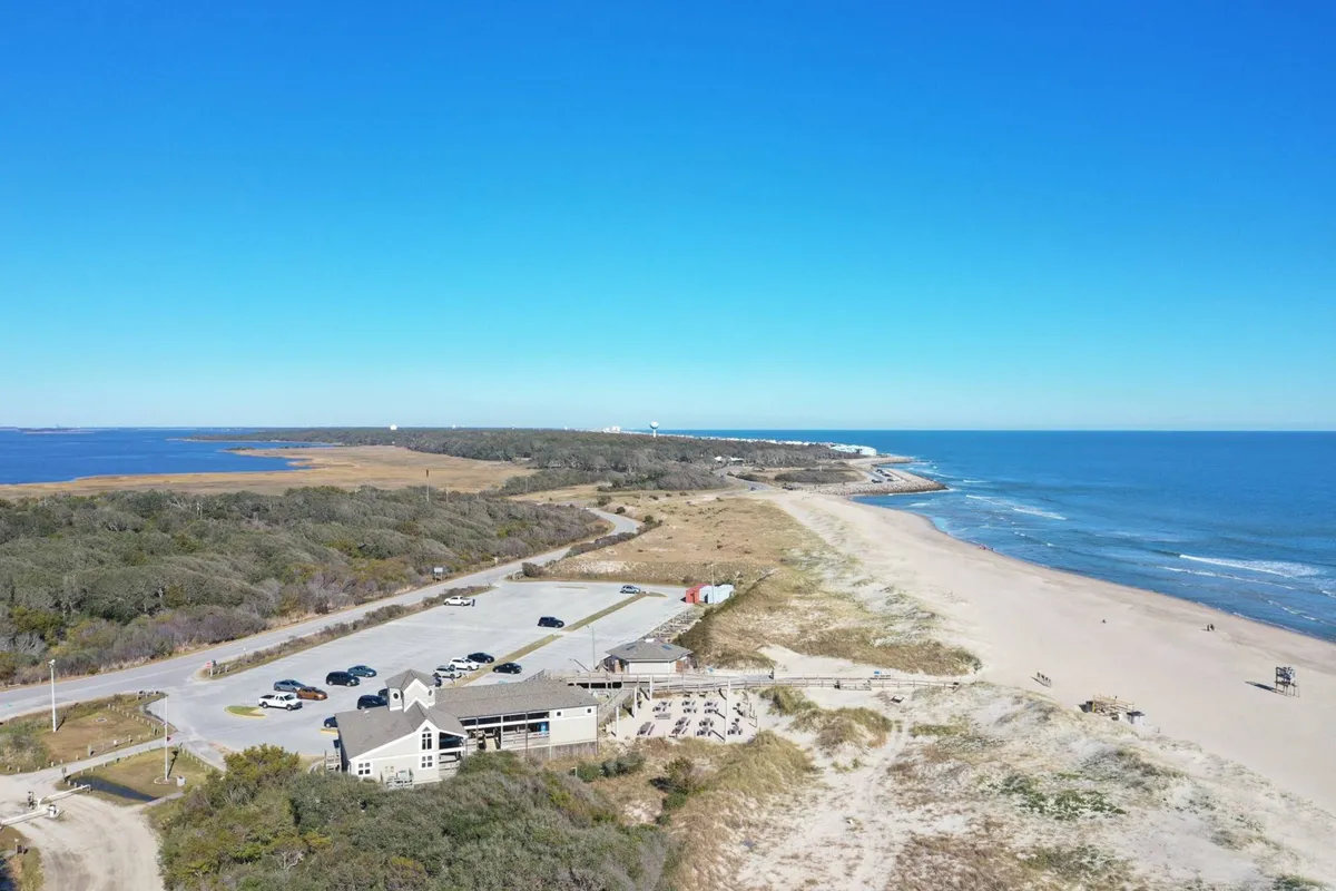 A wide view of the beach and ocean, with a visible parking area, a restaurant, and a winding path leading to the sand. The scene suggests easy access, local amenities, and opportunities for exploring the neighborhood’s food and nightlife.