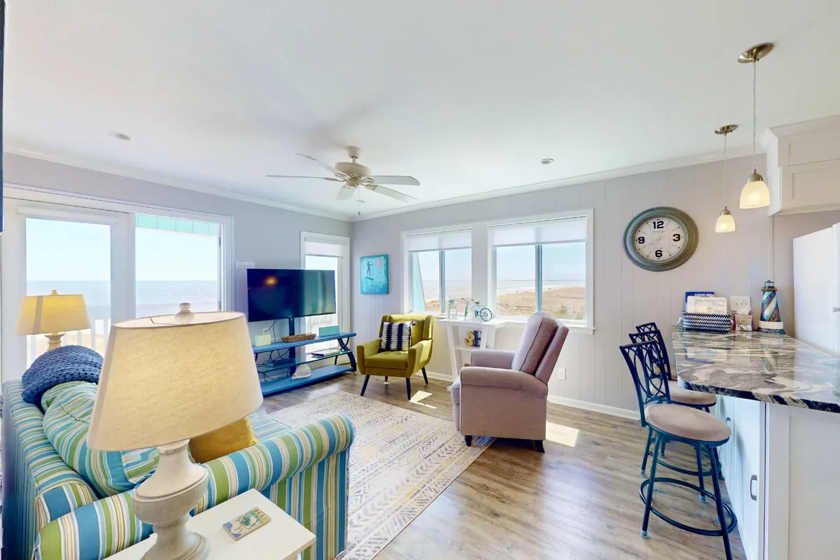 A spacious, inviting living area with a striped sofa, armchair, bar counter, and large windows framing a stunning ocean view. The room is filled with natural light and offers both relaxation and social areas, enhancing the open, airy feel of the top-floor vantage.