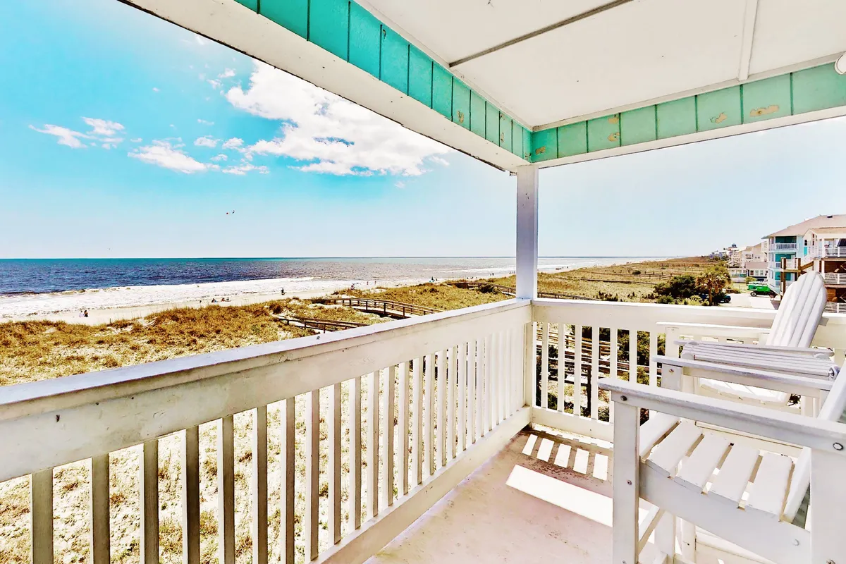 The image captures a serene balcony with two inviting white chairs overlooking the endless blue of the Atlantic Ocean. Puffy clouds drift across the vibrant sky, while the gentle waves and sandy shore beckon just below. The perspective promises panoramic ocean views and a tranquil escape.