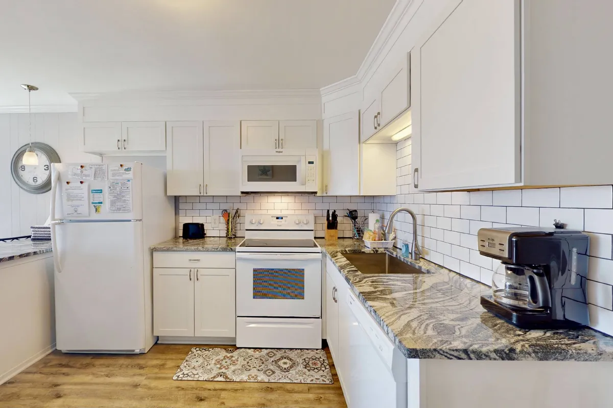A spacious, bright kitchen featuring granite countertops, white cabinetry, stainless steel appliances, and a decorative rug. The layout is both functional and stylish, with plenty of workspace for meal prep and all the essentials visible, including a coffee maker and toaster.