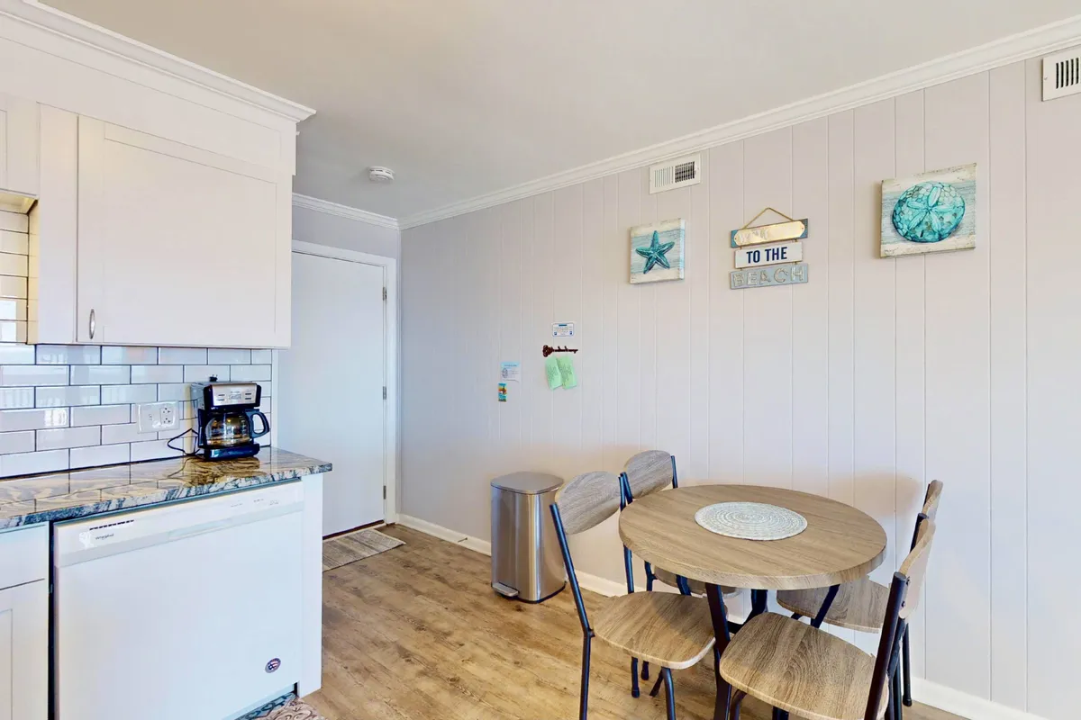 A cozy dining nook featuring a round table surrounded by comfortable chairs, complemented by a backdrop of kitchen appliances and decorative wall art that adds a welcoming touch. The scene is set for casual dining or morning coffee with family.