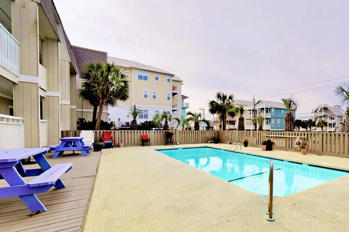 A vibrant exterior shot of the community swimming pool surrounded by lounge chairs, picnic tables, and palm trees. The pool area looks inviting, with ample space for guests to relax, swim, or socialize. The setting captures the essence of seasonal leisure and community gathering.
