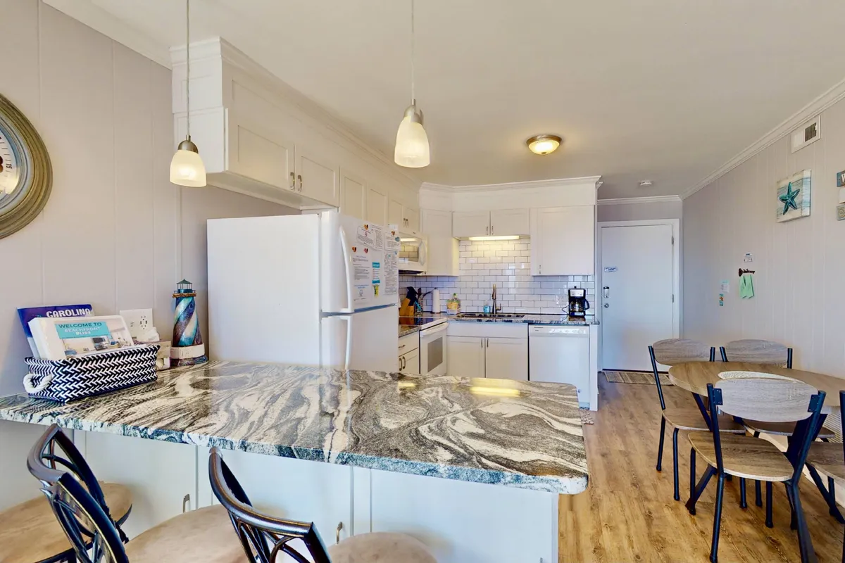 A cheerful open-concept kitchen and dining area with granite countertops, white cabinetry, modern pendant lighting, and a dining table ready for gatherings. The atmosphere is bright and social, perfect for both meals and group activities like board games.