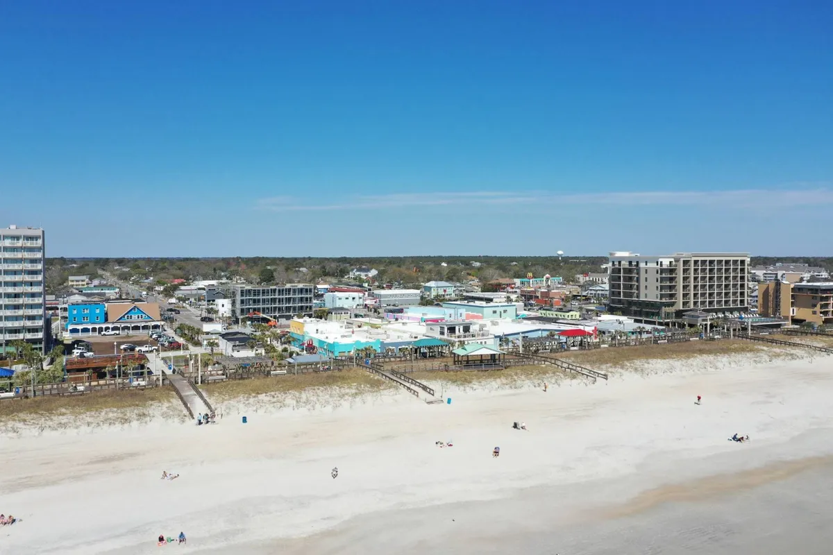 A lively exterior shot featuring the Carolina Beach boardwalk lined with colorful buildings and people enjoying the sun on the beach. The boardwalk’s proximity to the sand highlights easy access to dining, local shops, and festive activities.