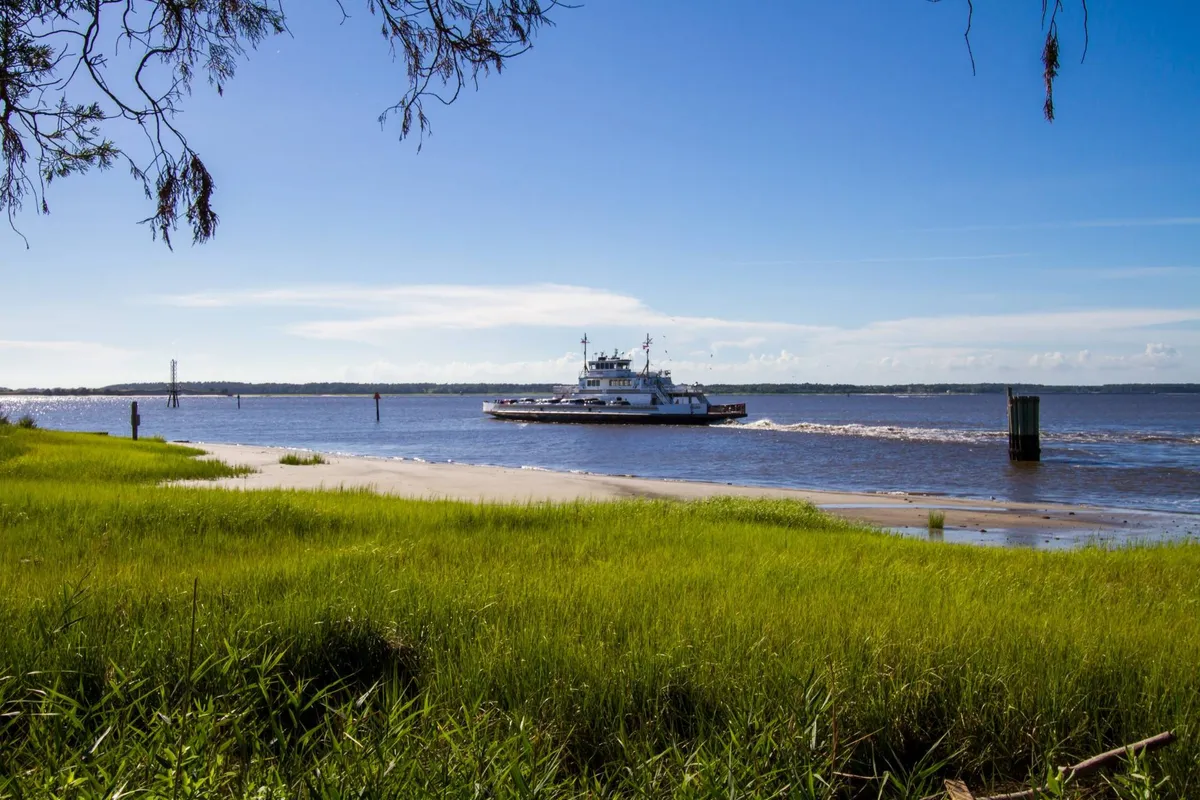 A serene view of a ferry gliding across a river under a blue sky, with green grass and calm waters in the foreground. The image hints at leisurely travel and local exploration opportunities just beyond the property.